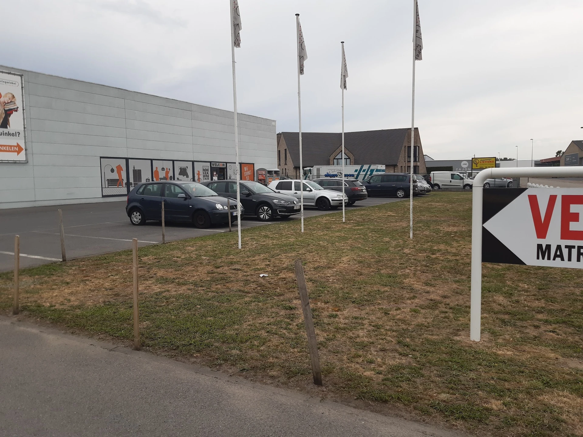 Parkeerterrein met auto's en vlaggen voor een grijze winkelgebouw
