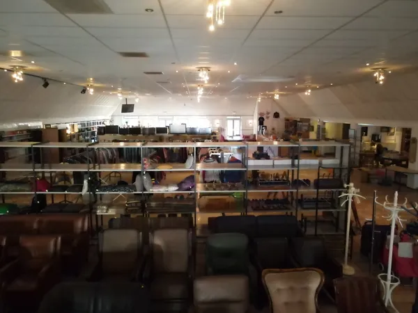 Overzicht van een kringloopwinkel met stellingen vol spullen, zitmeubelen op de vloer en verlichting aan het plafond