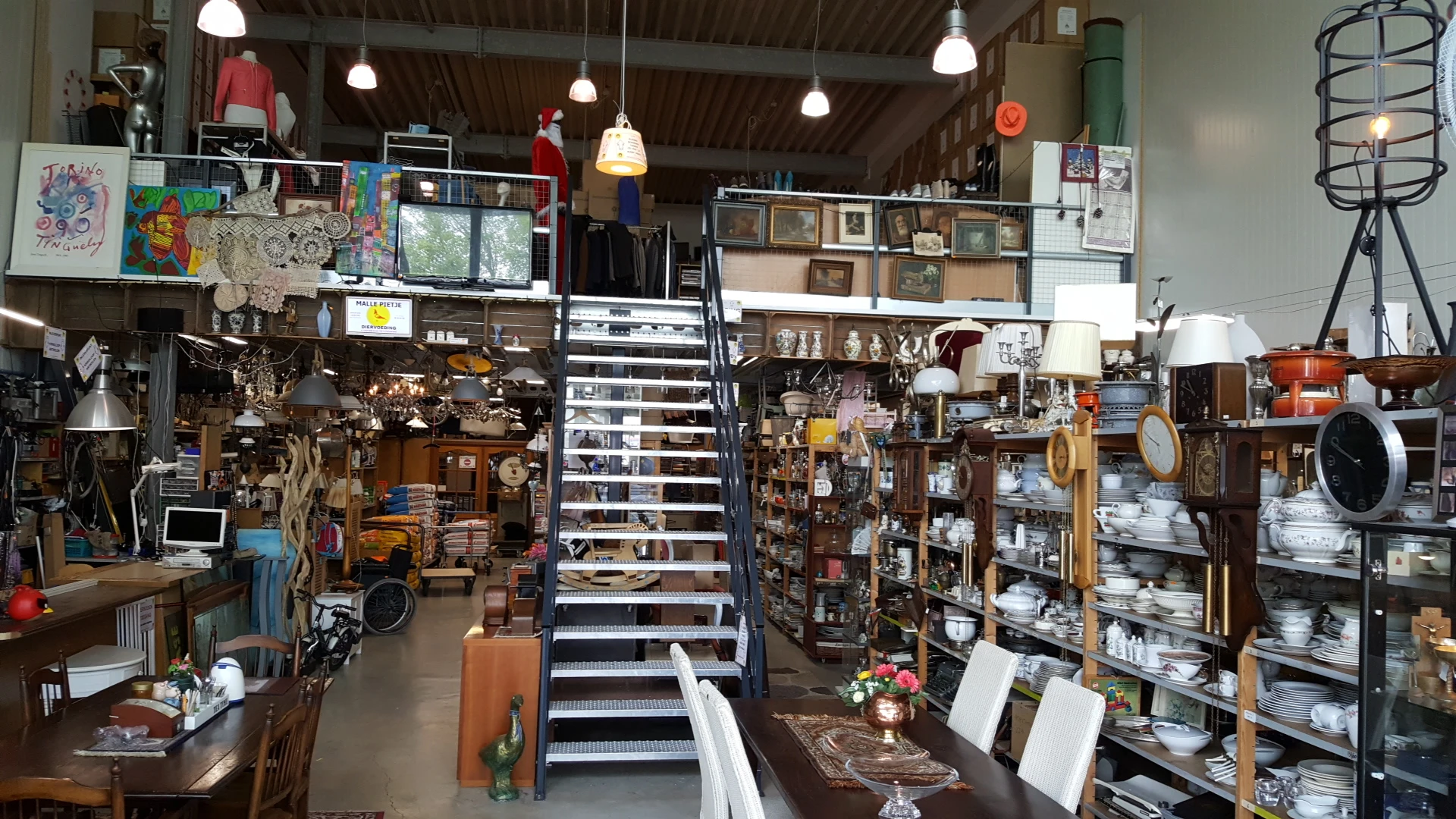 Overzicht van de kringloopwinkel met stellingen vol spullen, trappen in het midden en hangende lampen