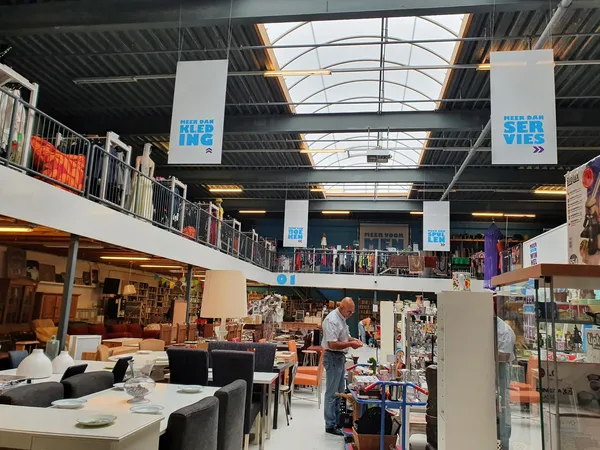 Interieur van kringloopwinkel met twee verdiepingen, stellingen met spullen, zithoek met stoelen en tafels, natuurlijk licht 