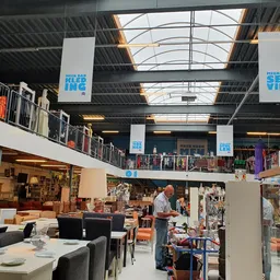 Interieur van kringloopwinkel met twee verdiepingen, stellingen met spullen, zithoek met stoelen en tafels, natuurlijk licht 