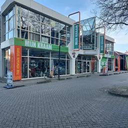 Gevel van kringloopwinkel Mijn Kraam met groene letters, winkelramen en glazen bovenbouw aan Noorder Passage in Emmen