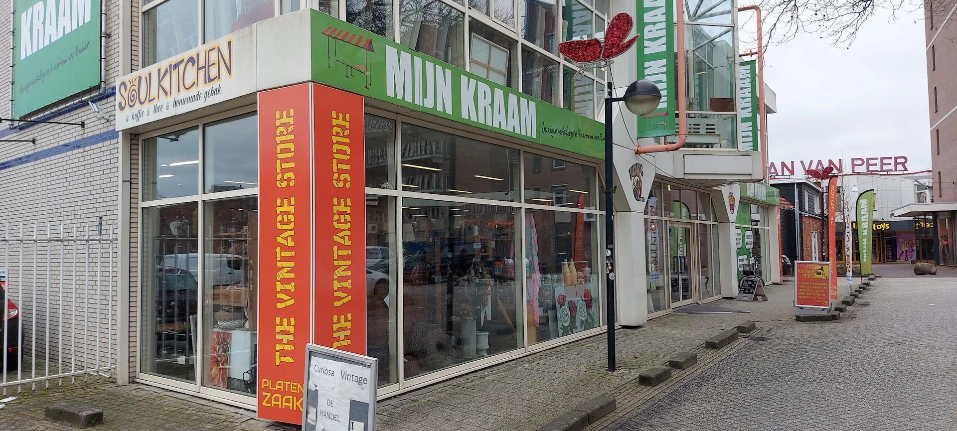 Gevel van kringloopwinkel met groene luifel, grote ramen en rode decoratieve letters aan de zijkant