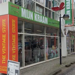 Gevel van kringloopwinkel met groene luifel, grote ramen en rode decoratieve letters aan de zijkant
