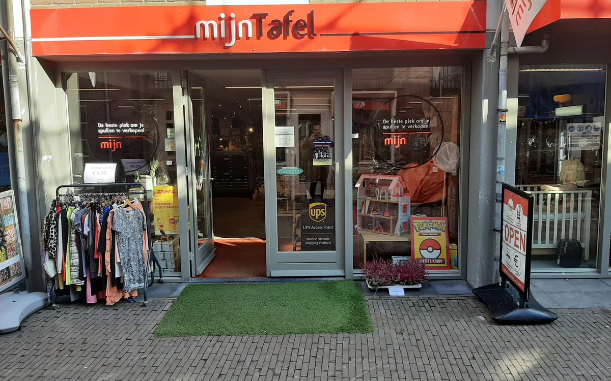 Winkelfront van mijn Tafel met rood luifel, etalageruiten met tweedehands kleding uitgestald buiten, groene deurmat
