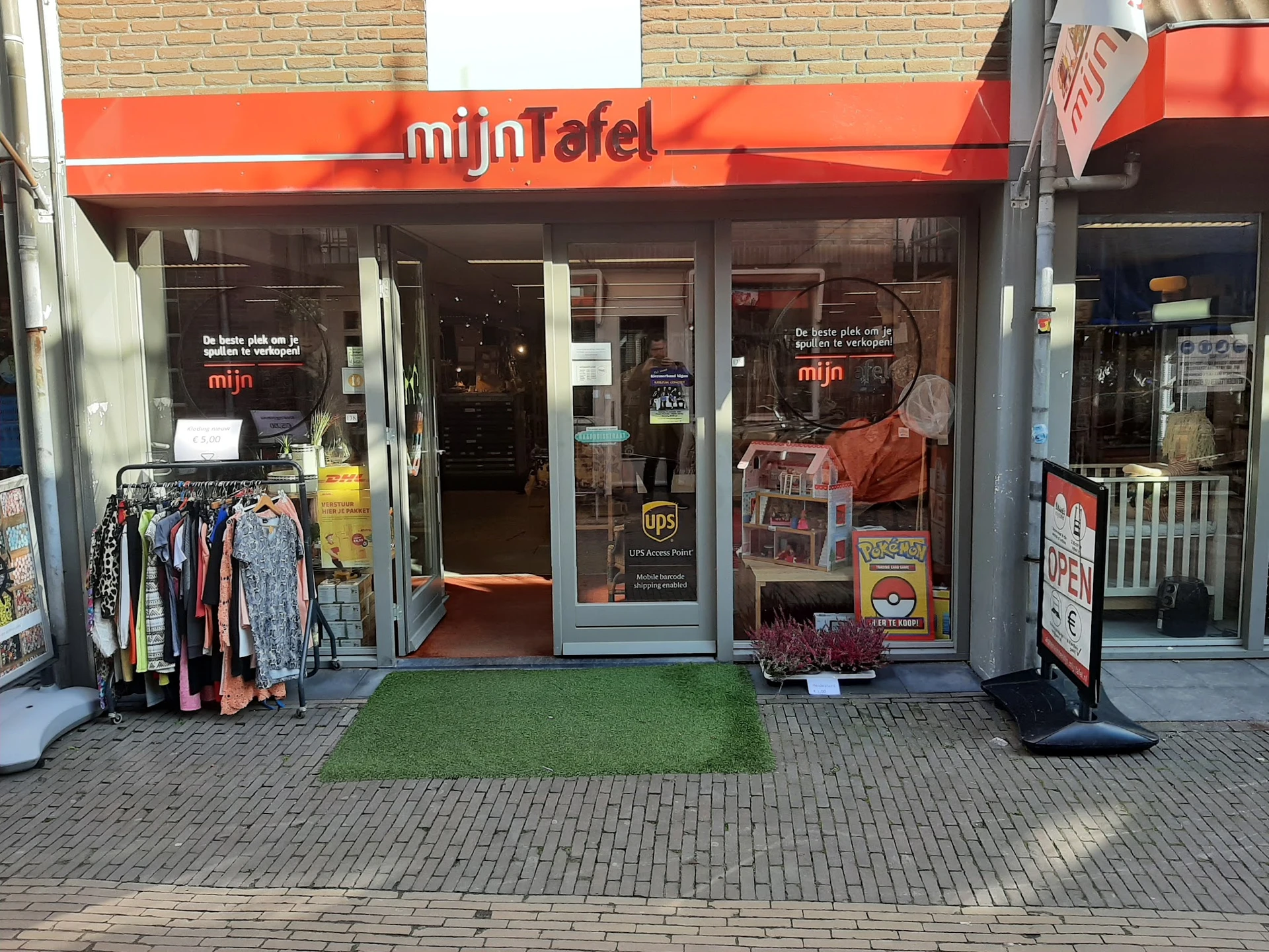 Winkelfront van mijn Tafel met rood luifel, etalageruiten met tweedehands kleding uitgestald buiten, groene deurmat