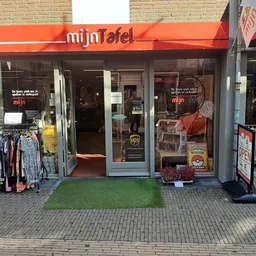 Winkelfront van mijn Tafel met rood luifel, etalageruiten met tweedehands kleding uitgestald buiten, groene deurmat