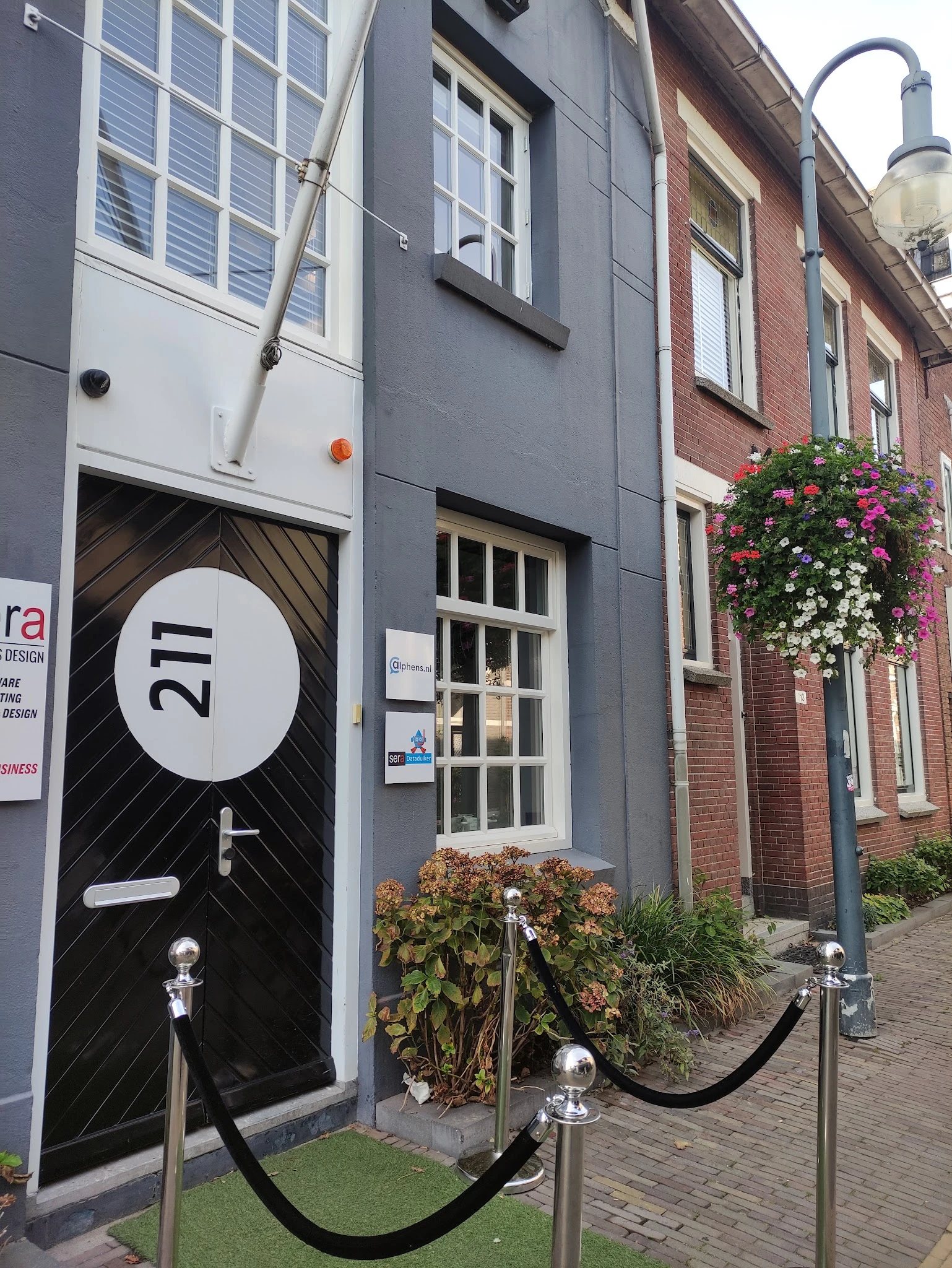 Gevel van een winkel aan de Raadhuisstraat met donkere deur genummerd 211, raam en bloembakken
