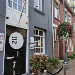 Gevel van een winkel aan de Raadhuisstraat met donkere deur genummerd 211, raam en bloembakken
