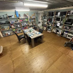 Winkelvloer met houten vloer, stellingen vol spullen aan beide zijden en een centrale tafel met witte stoelen in een kringloo