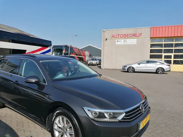 Parkeerterrein met zwarte auto op voorgrond, beige bedrijfsgebouw met 'AUTOBEDRIJF' opschrift en rode garagedeuren op achterg