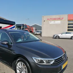 Parkeerterrein met zwarte auto op voorgrond, beige bedrijfsgebouw met 'AUTOBEDRIJF' opschrift en rode garagedeuren op achterg