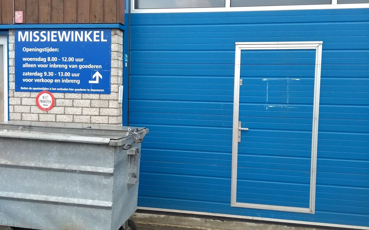 Blauwe gevel van kringloopwinkel met witte deur en informatiebord met openingstijden, grijze afvalcontainer ervoor