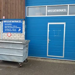 Blauwe gevel van kringloopwinkel met witte deur, openingstijden op bord, grijze afvalbak op straat
