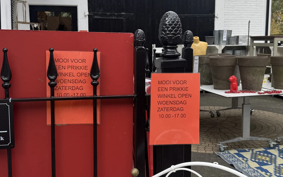 Voorkant van kringloopwinkel met rode panelen, zwarte deur en oranje openingsuren bord