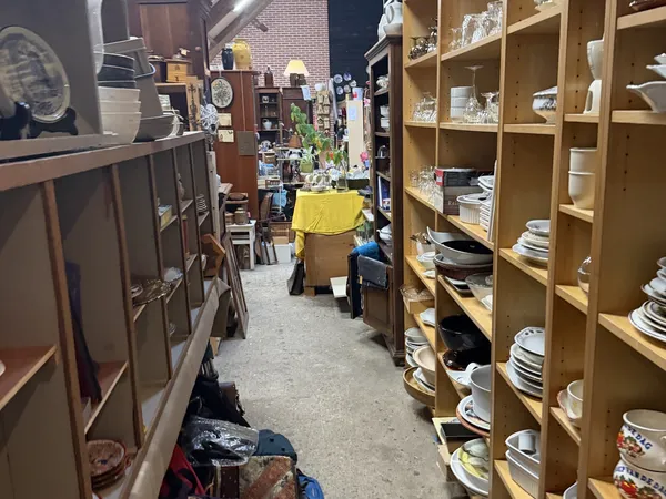 Winkelinterieur met houten stellingen vol serviesgoed en borden aan de rechterkant, donkere kasten links, gele doos op de vlo