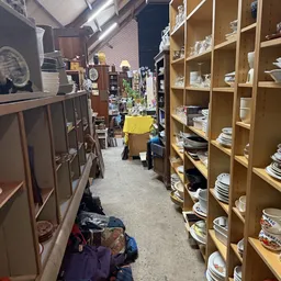 Winkelinterieur met houten stellingen vol serviesgoed en borden aan de rechterkant, donkere kasten links, gele doos op de vlo