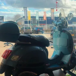 Blauwe Vespa-scooter met zwart zadel op een parkeerplaats met auto's en winkelcentrum op achtergrond