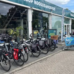 Voorgevel van kringloopwinkel met grote ramen, winkelnaam zichtbaar, fietsen en blauwe winkelwagen voor de ingang