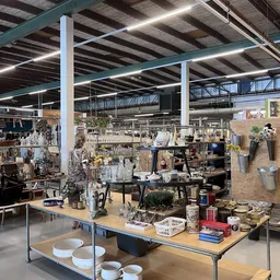 Werkplek in kringloopwinkel met tafels, stellingen, bloemen, decoratie en diverse spullen tussen hoge industriële plafonds