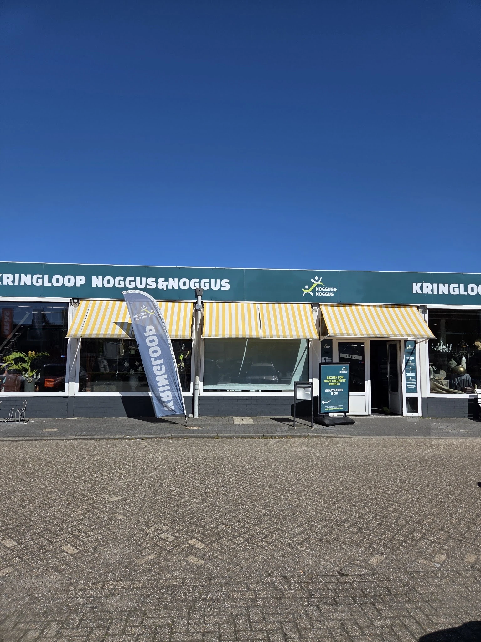Voorgevel van kringloopwinkel met geel-wit gestreepte luifels, groene balk met winkelnaam en logo.