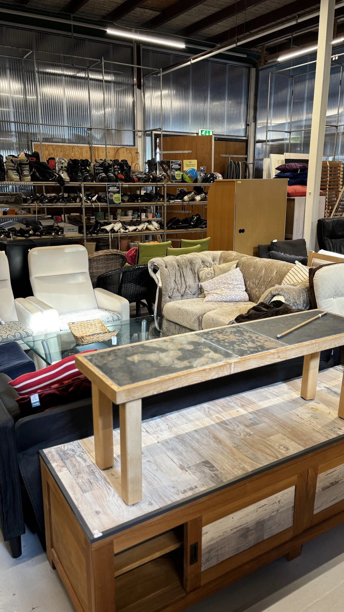 Meubels in de kringloopwinkel: een beige gestoffeerde bank met kussens, witte fauteuil en houten tafel met donkere blad