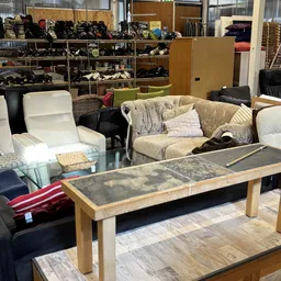 Meubels in de kringloopwinkel: een beige gestoffeerde bank met kussens, witte fauteuil en houten tafel met donkere blad