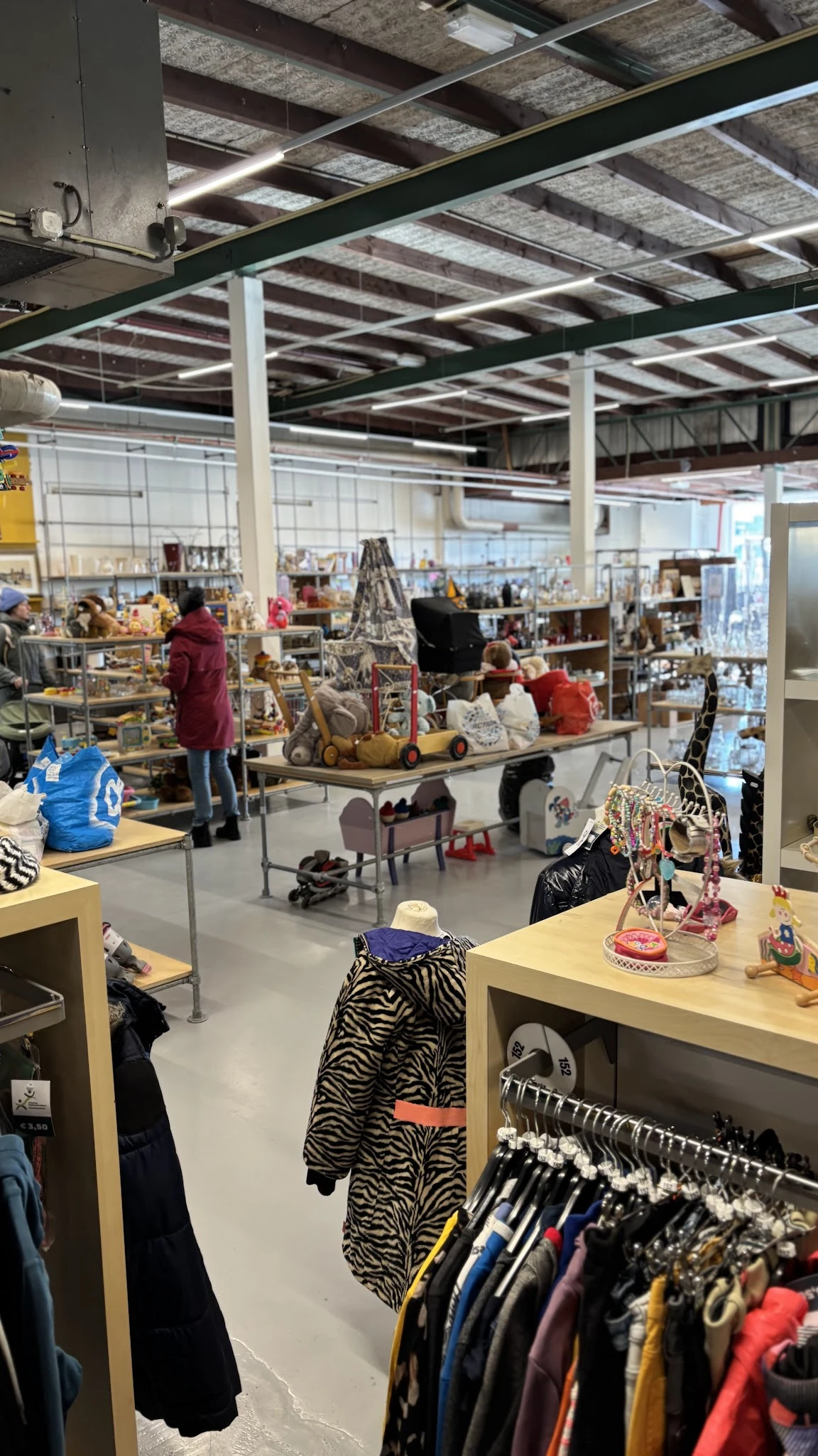 Winkelvloer van kringloopwinkel met verschillende afdelingen, tafels met spullen, klanten aan het winkelen, stellingen met ar