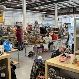 Winkelvloer van kringloopwinkel met verschillende afdelingen, tafels met spullen, klanten aan het winkelen, stellingen met ar