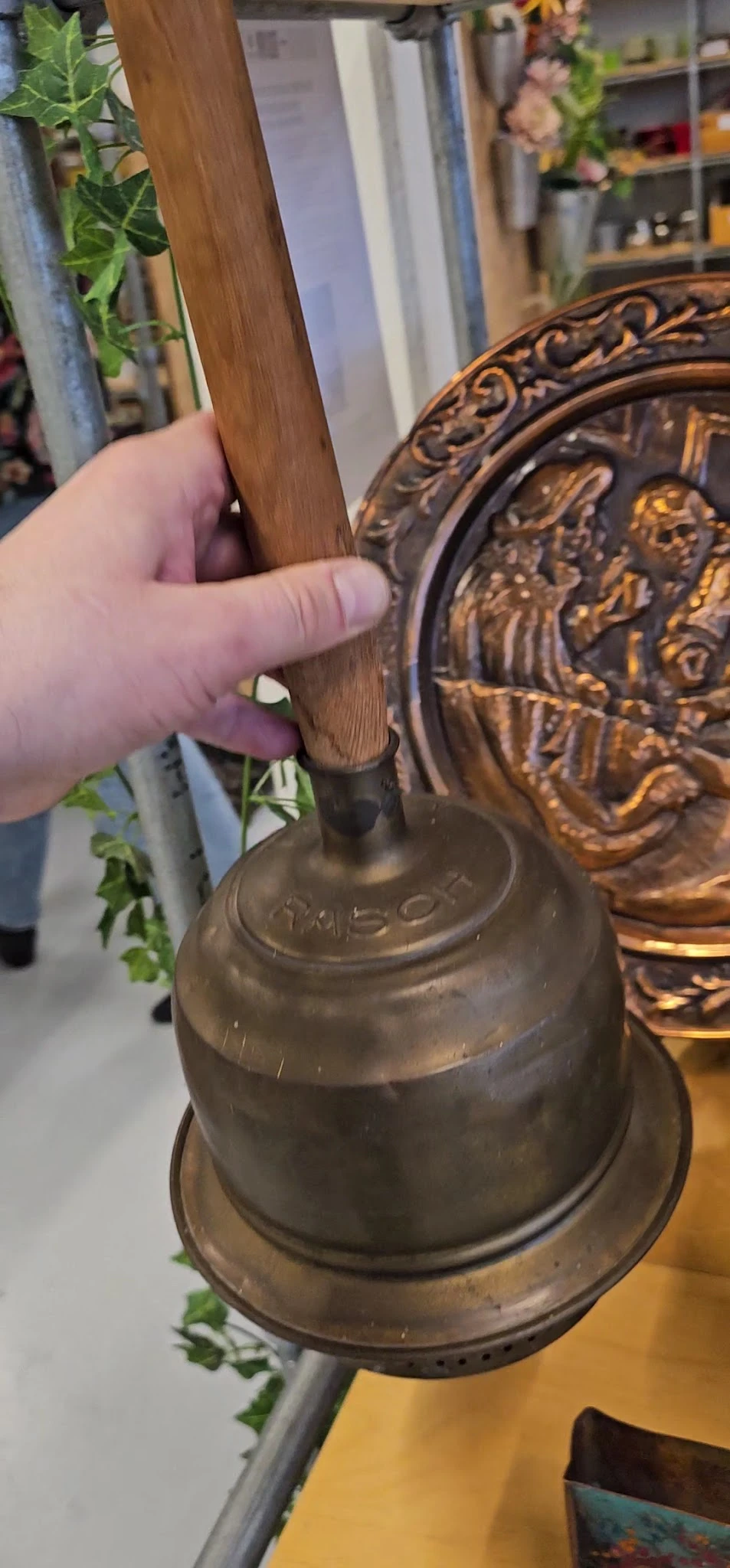Close-up van een bronzen bel met houten handvat, waarop iemand wijst naar het opschrift