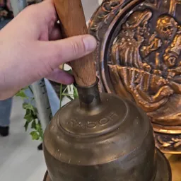 Close-up van een bronzen bel met houten handvat, waarop iemand wijst naar het opschrift
