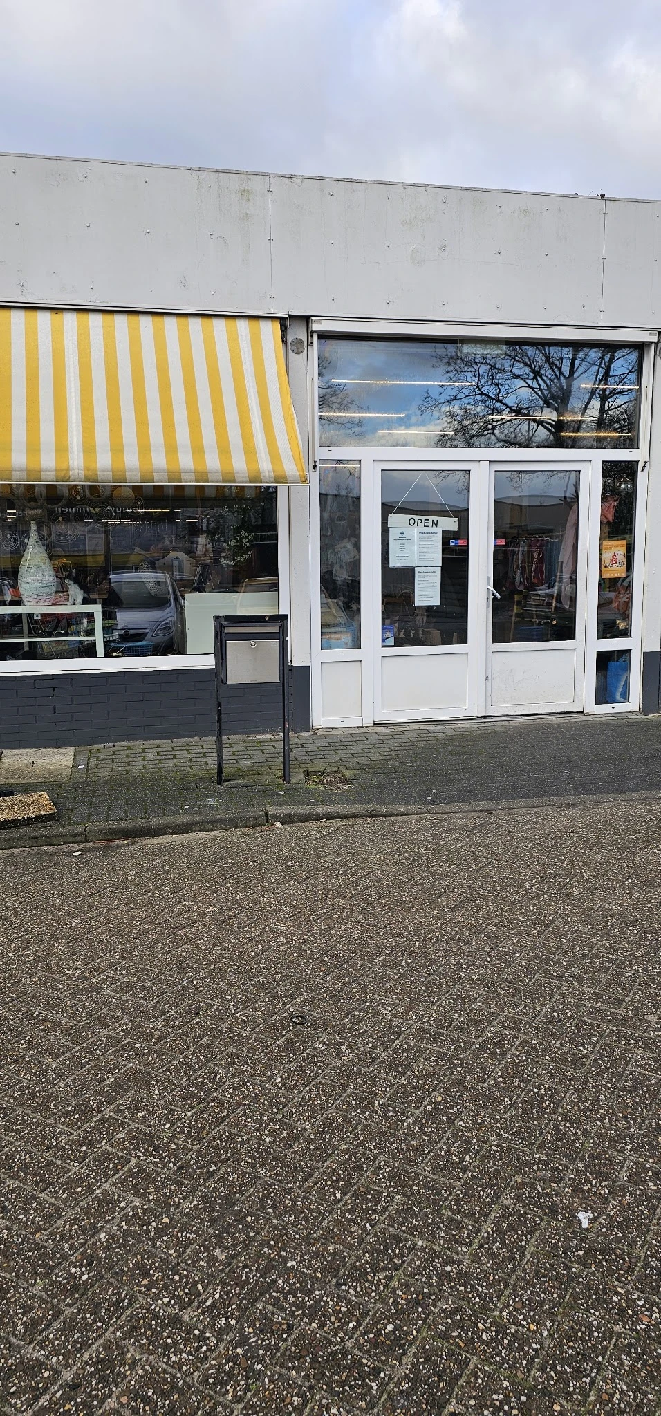 Voorgevel van kringloopwinkel met witte deuren en ramen, gedeeltelijk zichtbaar achter een afvalbak