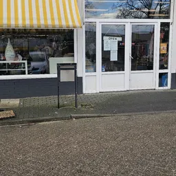 Voorgevel van kringloopwinkel met witte deuren en ramen, gedeeltelijk zichtbaar achter een afvalbak
