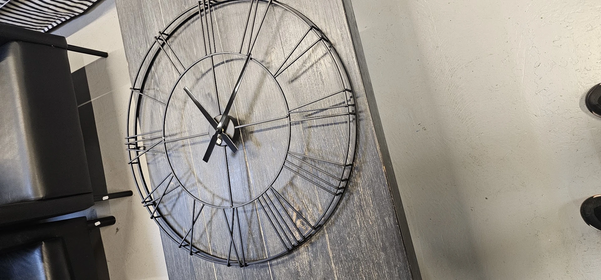 Metalen wandklok met open spijlwerk en zwarte wijzers tegen houten wand