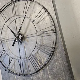 Metalen wandklok met open spijlwerk en zwarte wijzers tegen houten wand