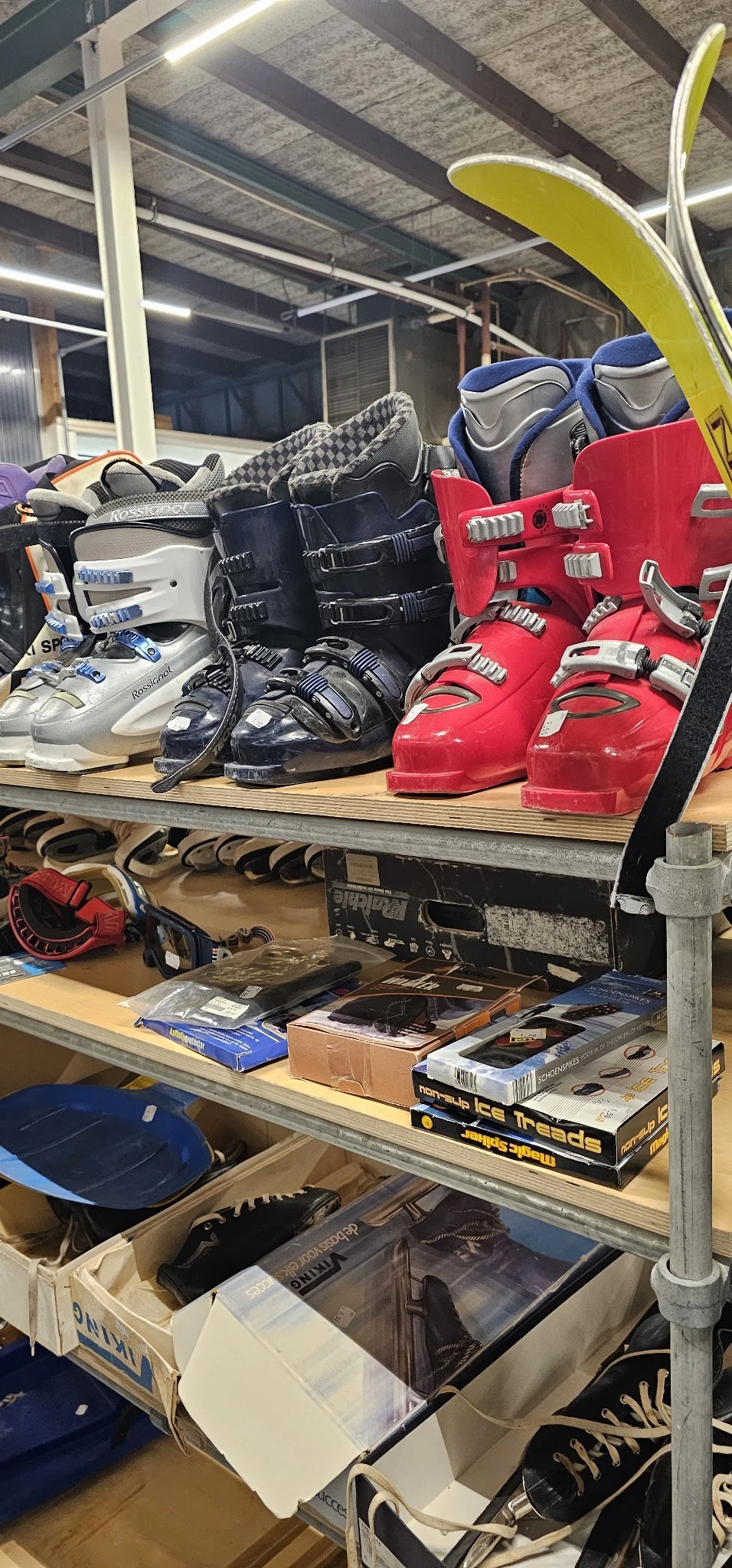 Verzameling tweedehands skischoenen in verschillende kleuren op een houten schap met accessoires