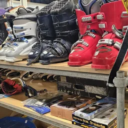 Verzameling tweedehands skischoenen in verschillende kleuren op een houten schap met accessoires