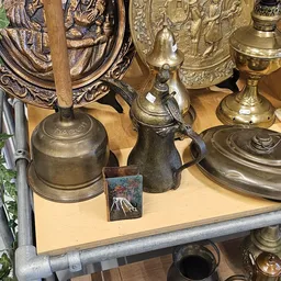 Uitgestalde antieke en vintage items: koperen en zilveren bellen, kannen, kelken en decoratieve objecten op houten plank