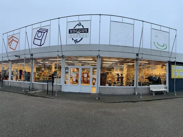 Gevel van kringloopwinkel met brede glazen pui, winkelnaam Noggus & Noggus zichtbaar, winkelramen met verlicht interieur