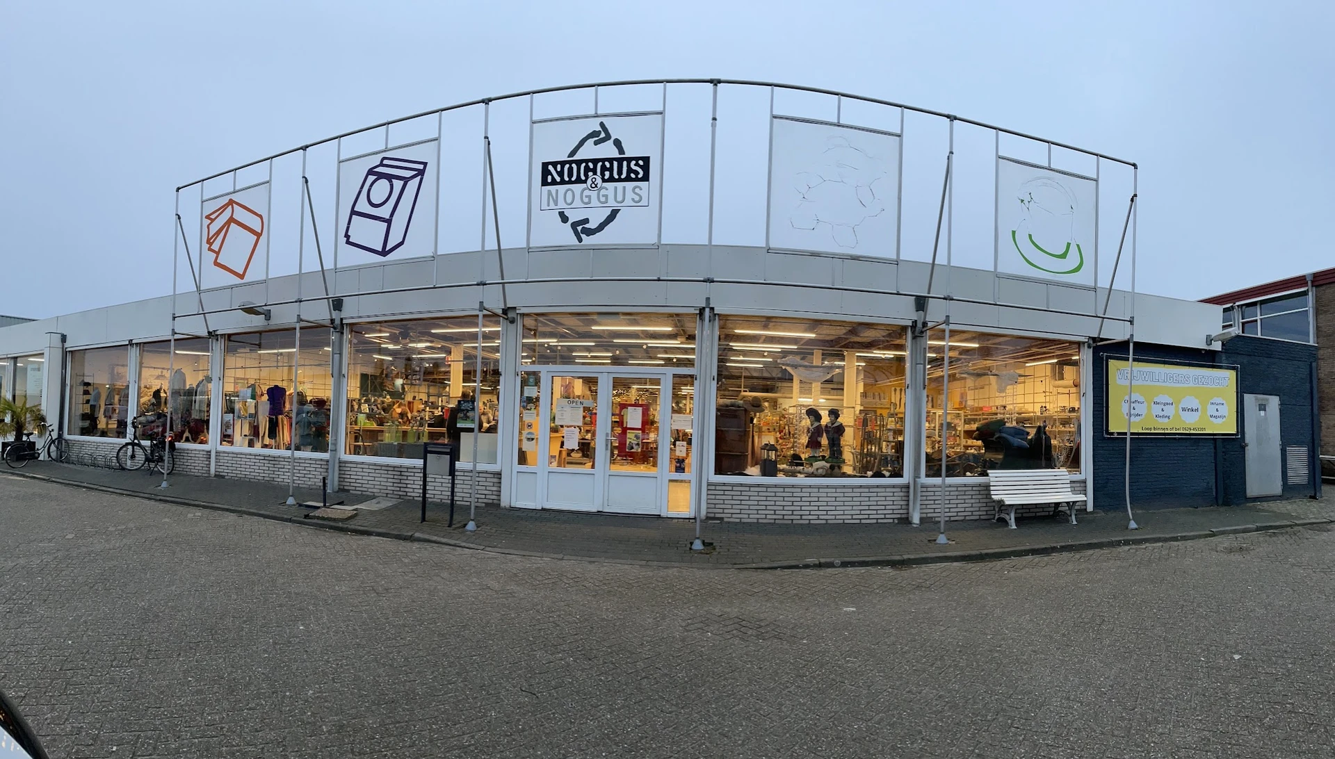 Gevel van kringloopwinkel met brede glazen pui, winkelnaam Noggus & Noggus zichtbaar, winkelramen met verlicht interieur