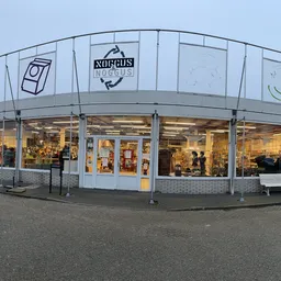 Gevel van kringloopwinkel met brede glazen pui, winkelnaam Noggus & Noggus zichtbaar, winkelramen met verlicht interieur