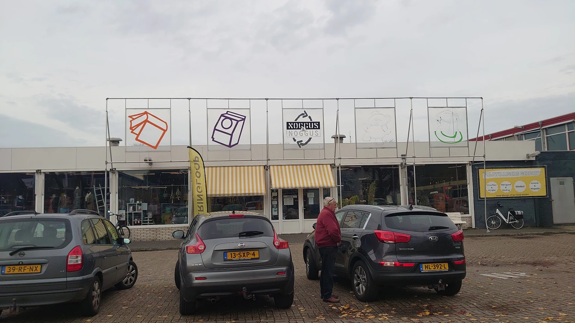Gevelfront van kringloopwinkel met witte pui, gekleurde logo's en parkeerplaats met auto's ervoor