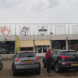 Gevelfront van kringloopwinkel met witte pui, gekleurde logo's en parkeerplaats met auto's ervoor