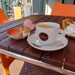 Kopje koffie met AGUST-logo op houten tafel met ijsje en gebak op terras