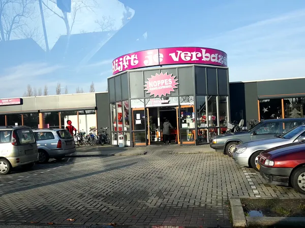 Exterieur van kringloopwinkel met roze cirkelvormig logo en parkeerplaats met auto's