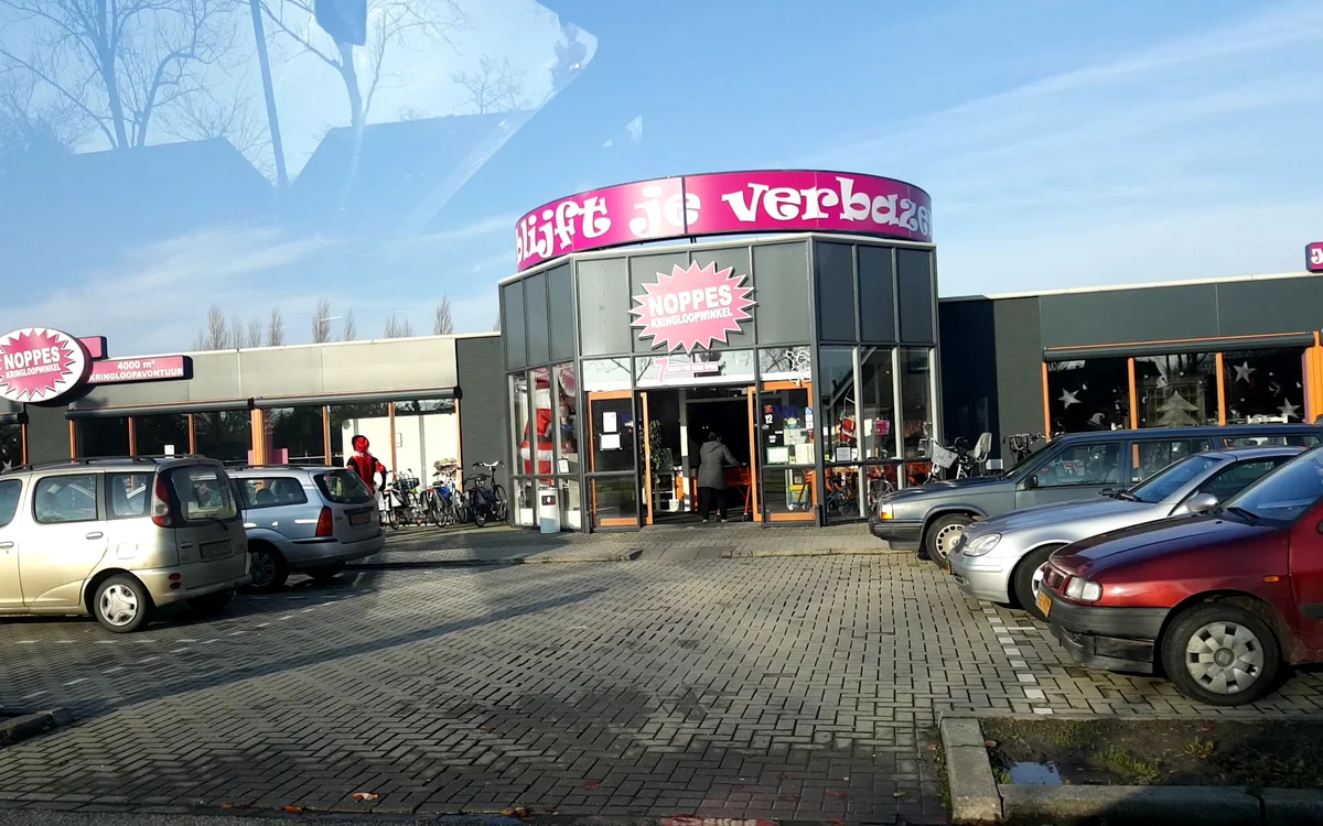 Exterieur van kringloopwinkel met roze cirkelvormig logo en parkeerplaats met auto's