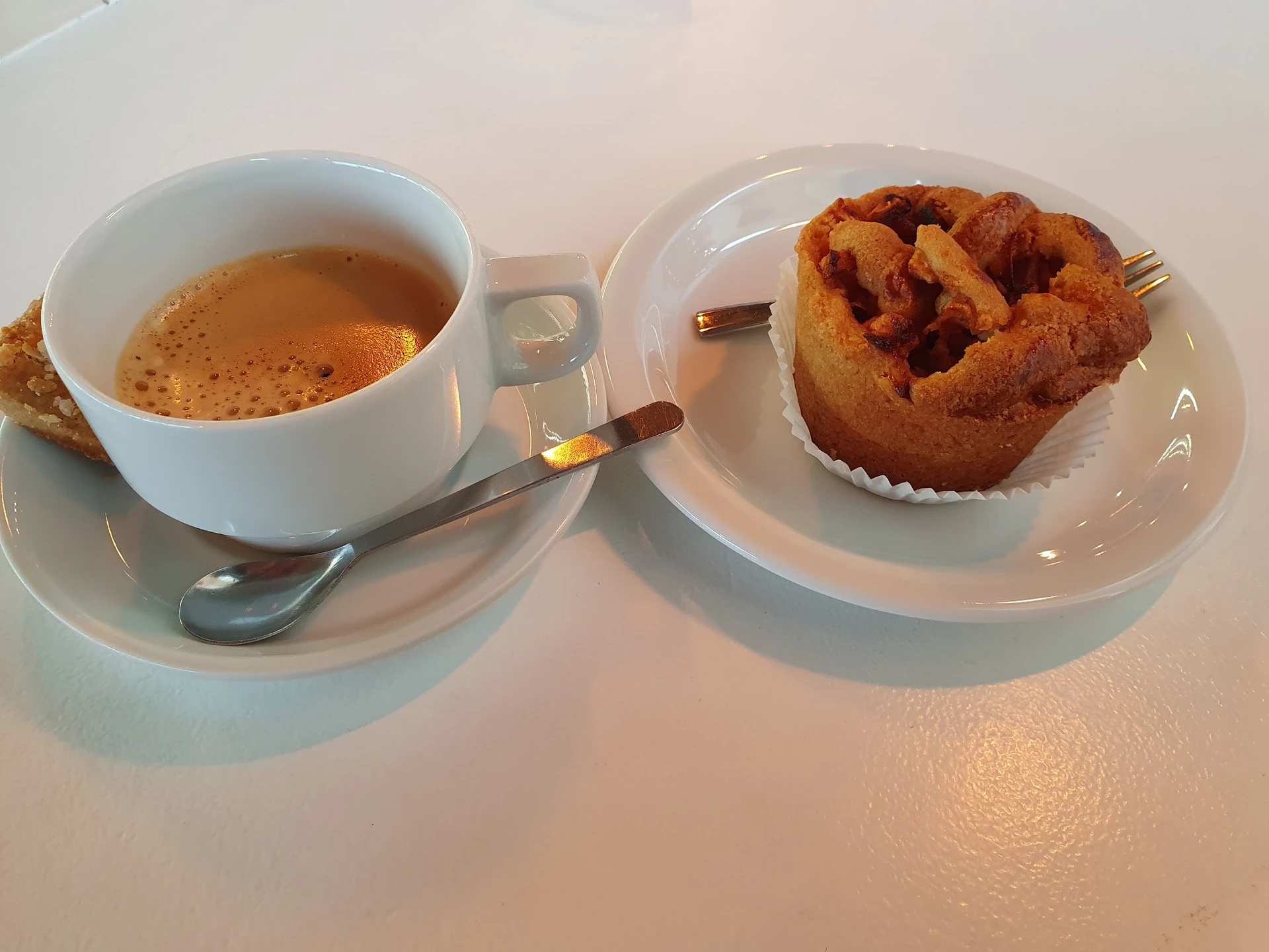Kopje koffie met schuim en gebakje op wit bord met lepel op wit schoteltje