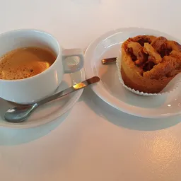 Kopje koffie met schuim en gebakje op wit bord met lepel op wit schoteltje