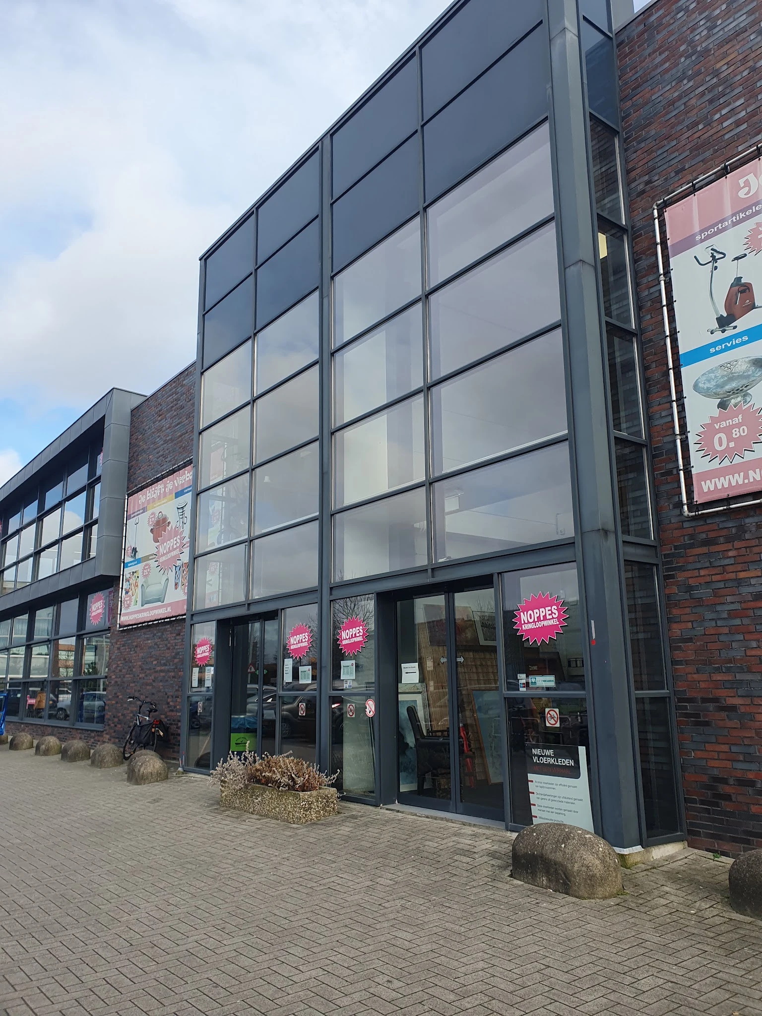 Voorgevel van kringloopwinkel met grote ramen, donkerblauwe pui en roze borden met 'NOPPES' letters