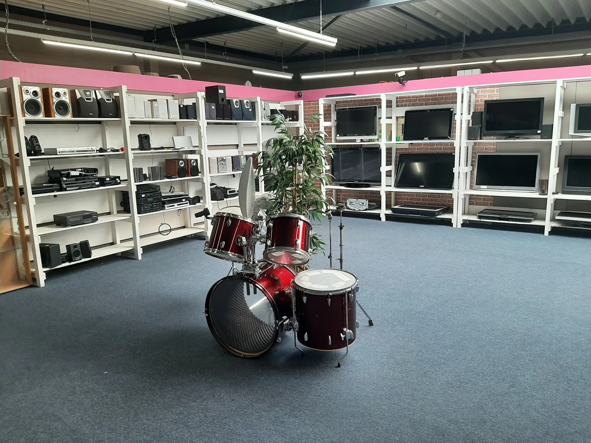 Interieur van kringloopwinkel met witte stellingen vol elektronische apparatuur, drums en een plant op de vloer